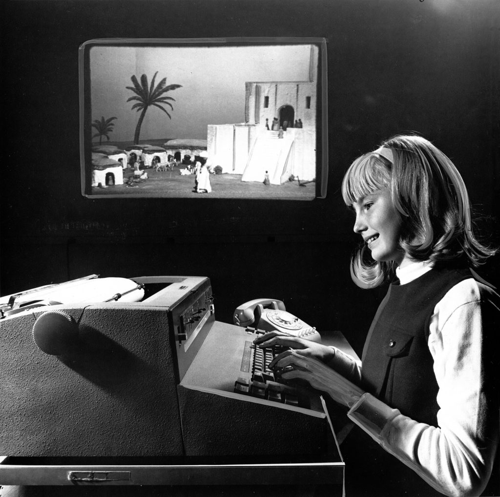 An unnamed girl sitting at a large typewriter playing The Sumerian Game with a slide projection screen in the background
